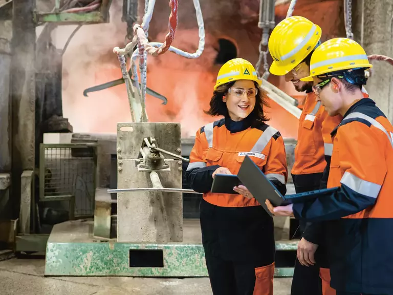 Zwei Männer und eine Frau bei der Arbeit in orangener Sicherheitskleidung mit gelben Helmen, Schutzbrillen und Mappen in den Händen, in einer Besprechung.