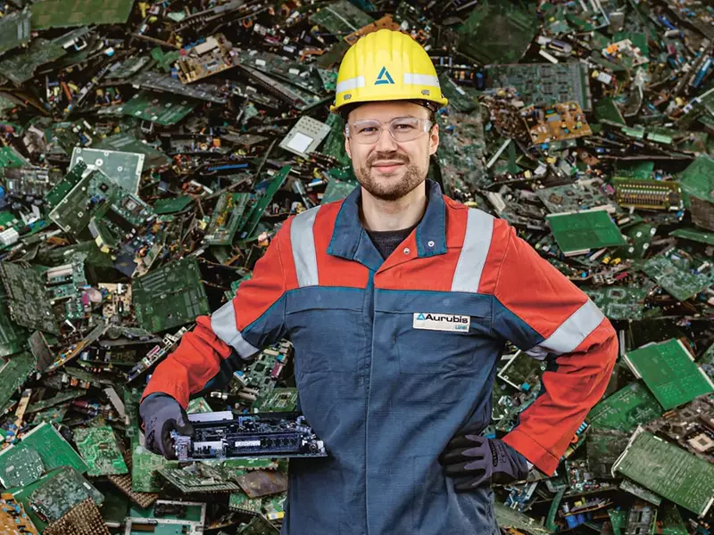 Tobias Kuhm, Executive Director Supply Chain Management bei Aurubis Lünen, der in Arbeitskleidung, Schutzbrille und Helm vor einem Berg an wiederverwertbarem Metallschrott steht.