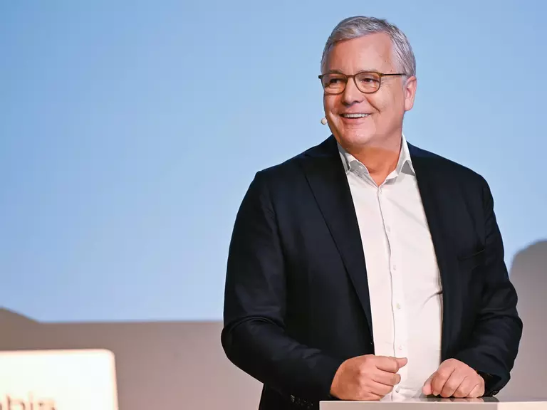 Dr. Toralf Haag am Rednerpult in schwarzem Anzug mit weißem Hemd, Brille und Namensschild vor hellblauem Hintergrund
