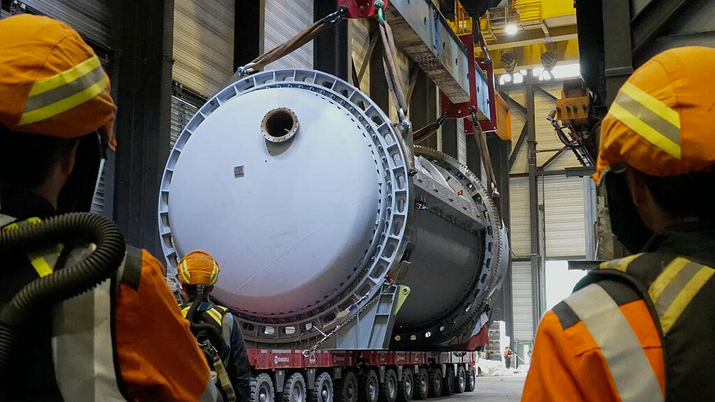 Zu sehen sind zwei Werksarbeitern in Schutzkleidung von hinten, die in einer Werkhalle vor einer Kranvorrichtung stehen, die eine auf Rollen bewegte Metallröhre zieht.