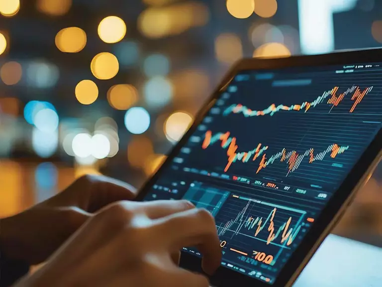 Im Fokus ein schwarzer Bildschirm, auf dem grüne und rote Graphen zu sehen sind, darunter tippende Hände, der Hintergrund verschwommen