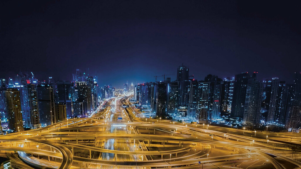 Skyline einer Großstadt bei Nacht, im Vordergrund ein leeres, von warmem Laternenschein beleuchtetes Straßennetzwerk