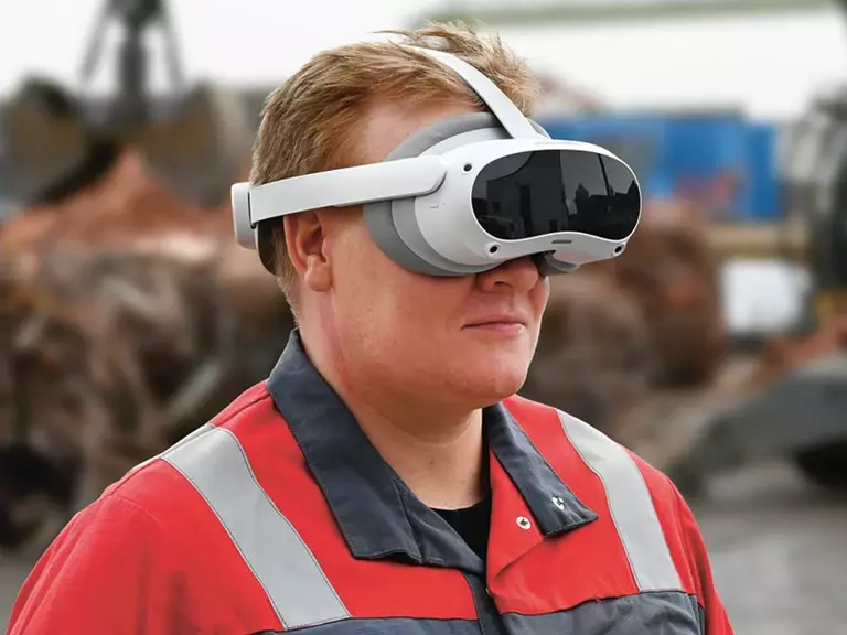 Mann mit Arbeitskleidung und VR-Brille auf einer Baustelle, im Hintergrund eine Kranschaufel