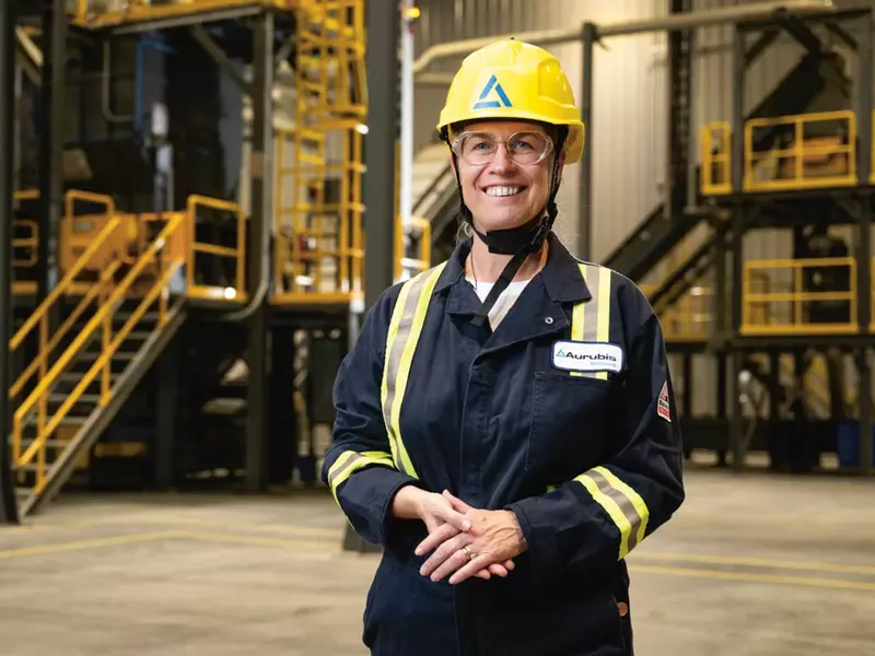 Inge Hofkens, COO, die mit gelbem Schutzhelm und dunkelblauer Arbeitskleidung mit Reflektoren in einer Arbeitshalle steht