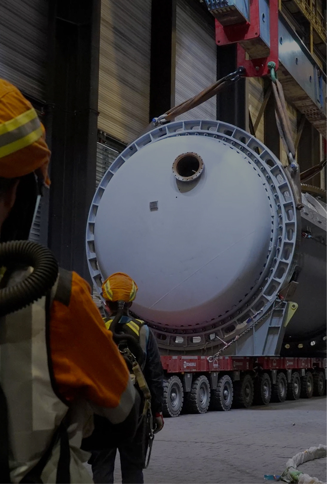 Erste Ebene der Aurubis Performance 2030: Impact. Zu sehen sind zwei Werksarbeitern in Schutzkleidung von hinten, die in einer Werkhalle vor einer Kranvorrichtung stehen, die eine auf Rollen bewegte Metallröhre zieht.